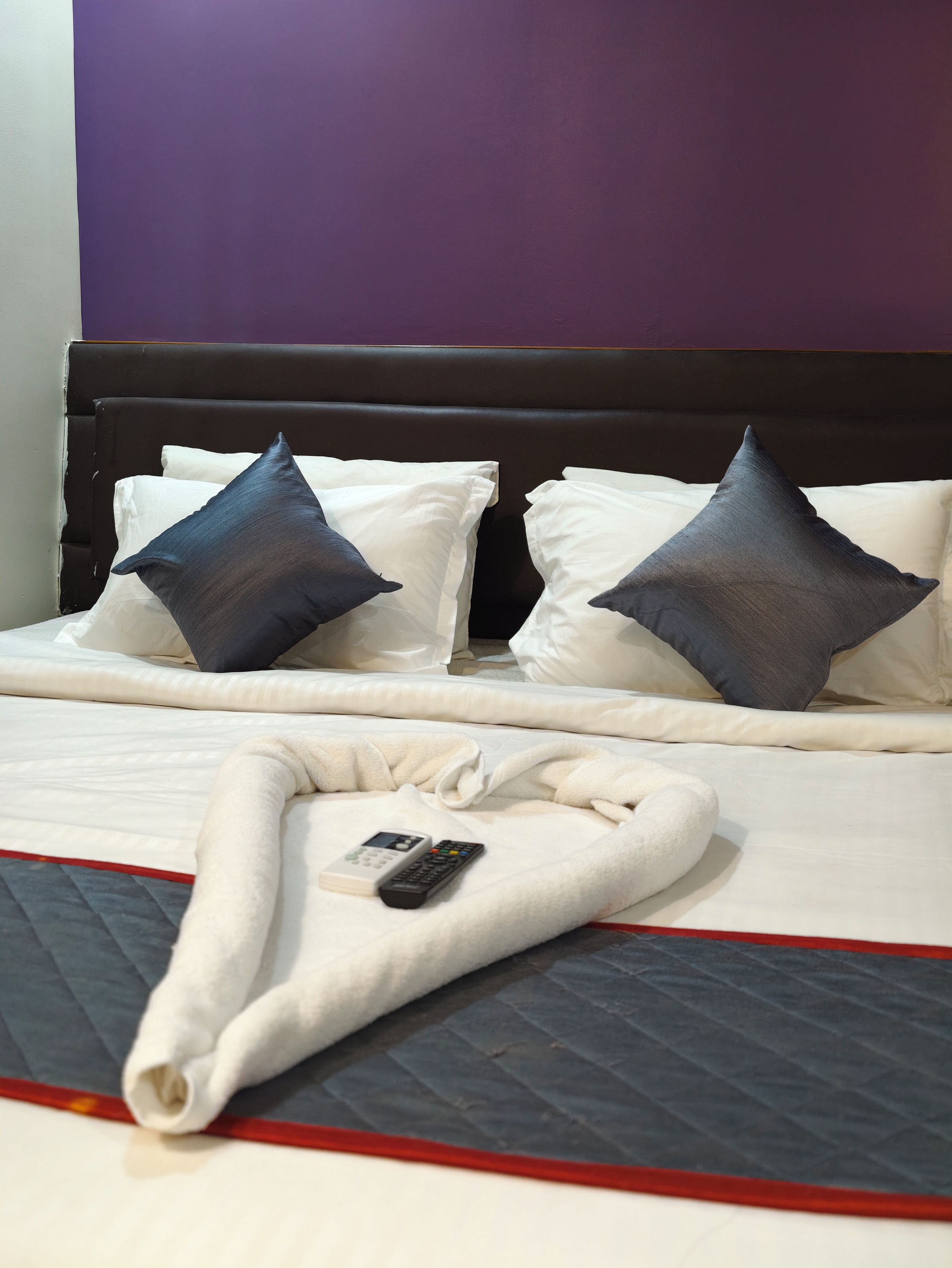 A neatly made bed with white and dark pillows, a remote control, and a folded blanket on a bed with a red trim.