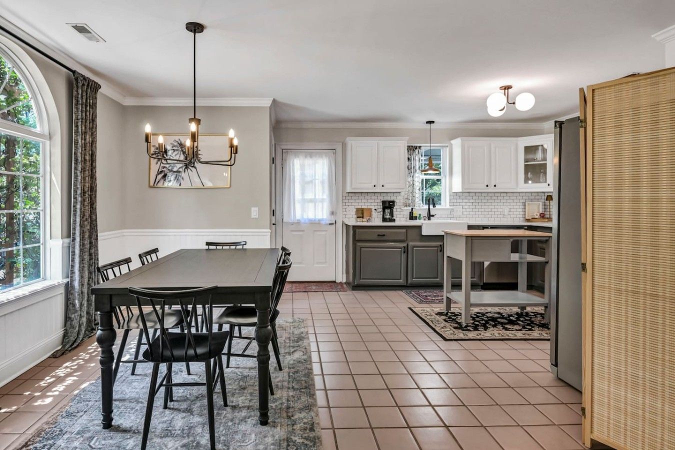 A dining area with a table and chairs, a kitchen with white cabinets and a tiled floor, and a large window with curtains.