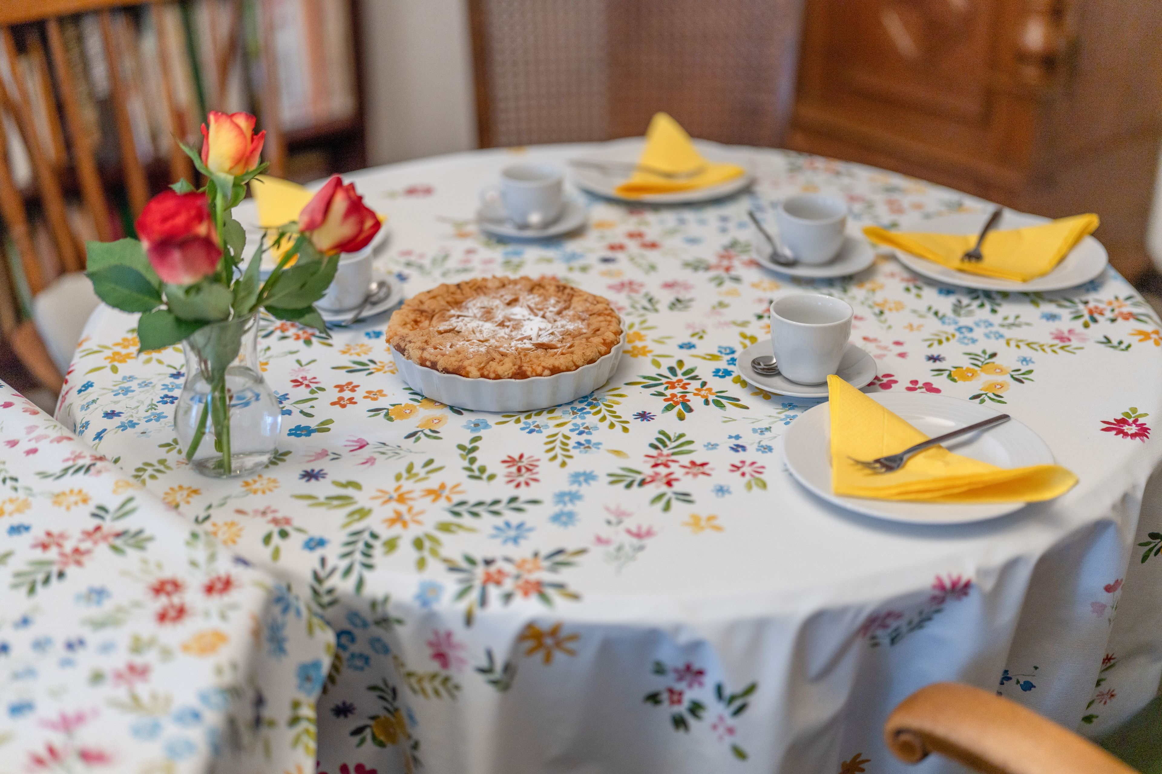 Une table ronde recouverte d’une nappe à motifs floraux, une tarte, des tasses et des serviettes jaunes.