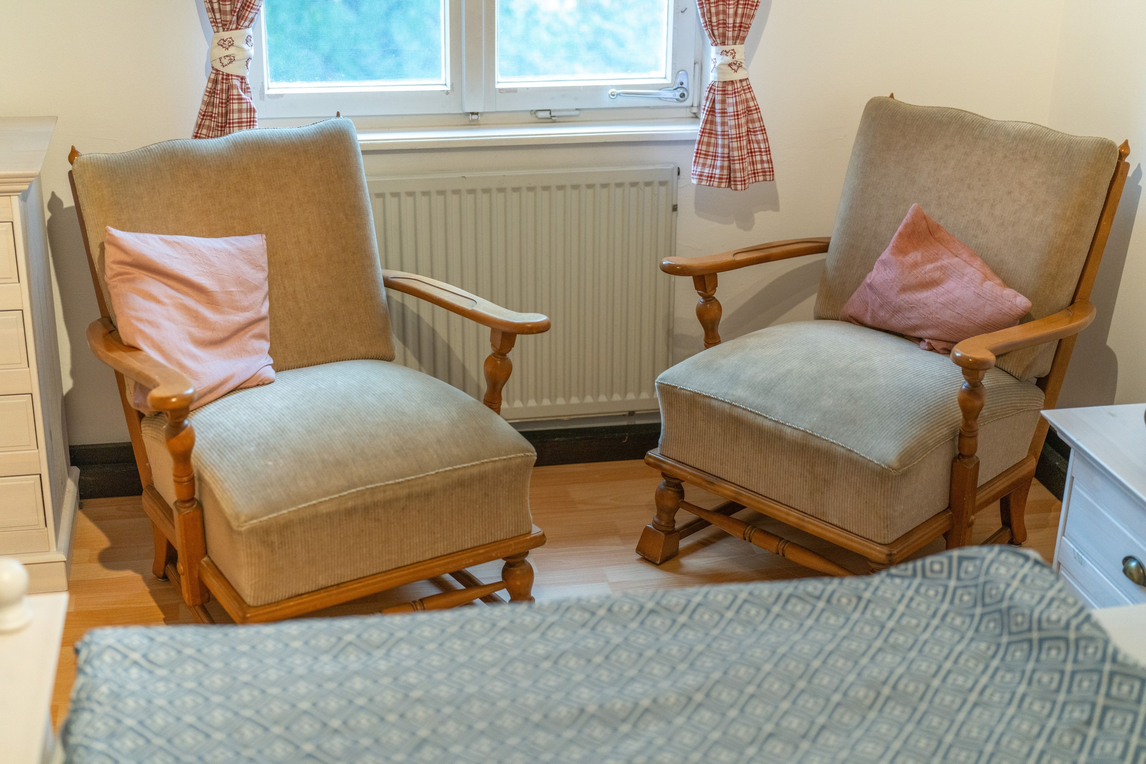 Une pièce comprenant deux fauteuils en bois, un radiateur, une fenêtre avec des rideaux et un lit recouvert d’une couvre-lit à motifs.