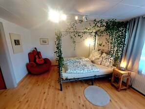 A bedroom with a bed, a guitar, a red chair, a bedside table with a lamp, and a radiator.