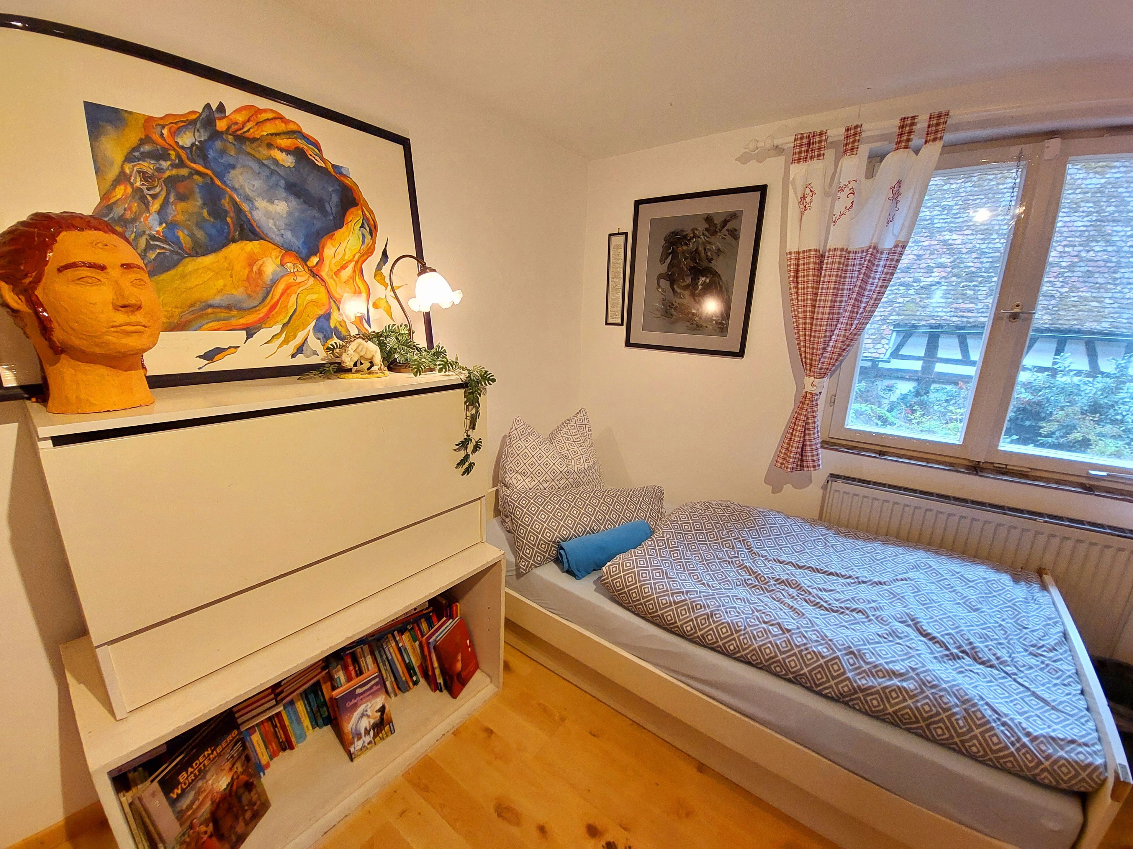 A bedroom with a bed, a bookshelf, a window with curtains, and a painting on the wall.