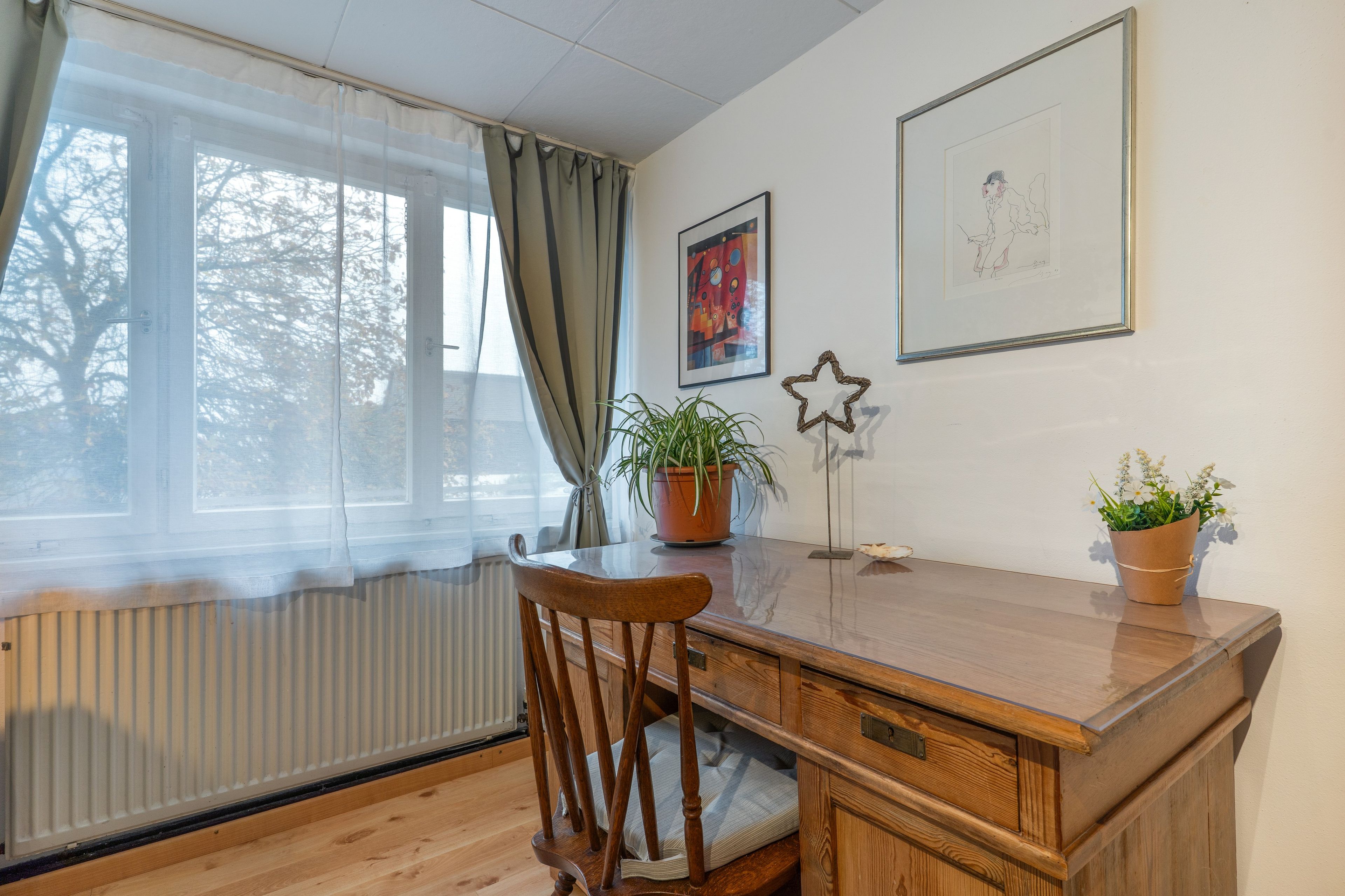 Een kamer met een houten bureau, een stoel, een radiator, een raam met gordijnen en twee ingekaderde kunstwerken aan de muur.