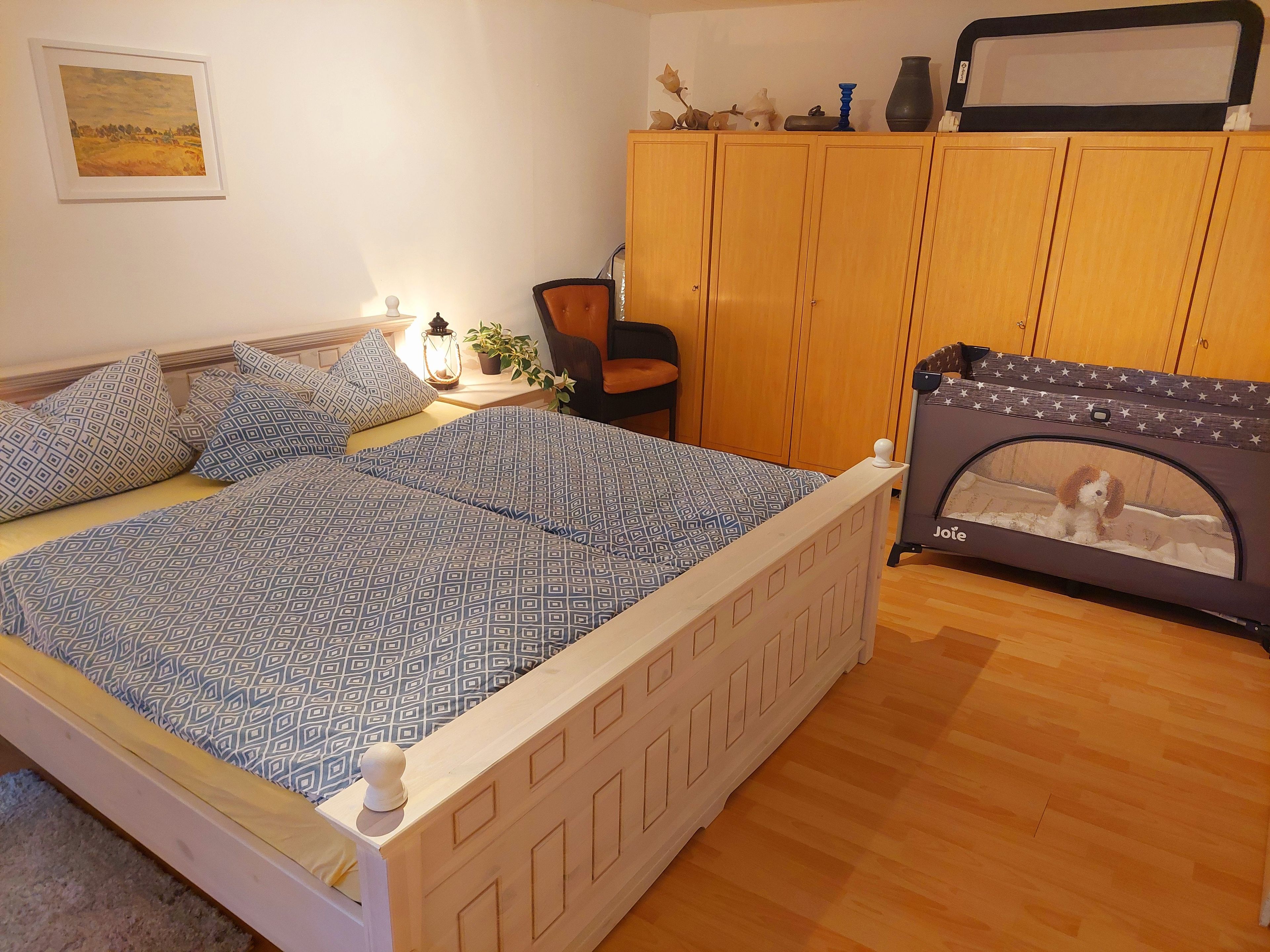 Une chambre à coucher comprenant un lit, une table de chevet, une lampe, une chaise, une armoire et un panier pour animal de compagnie.