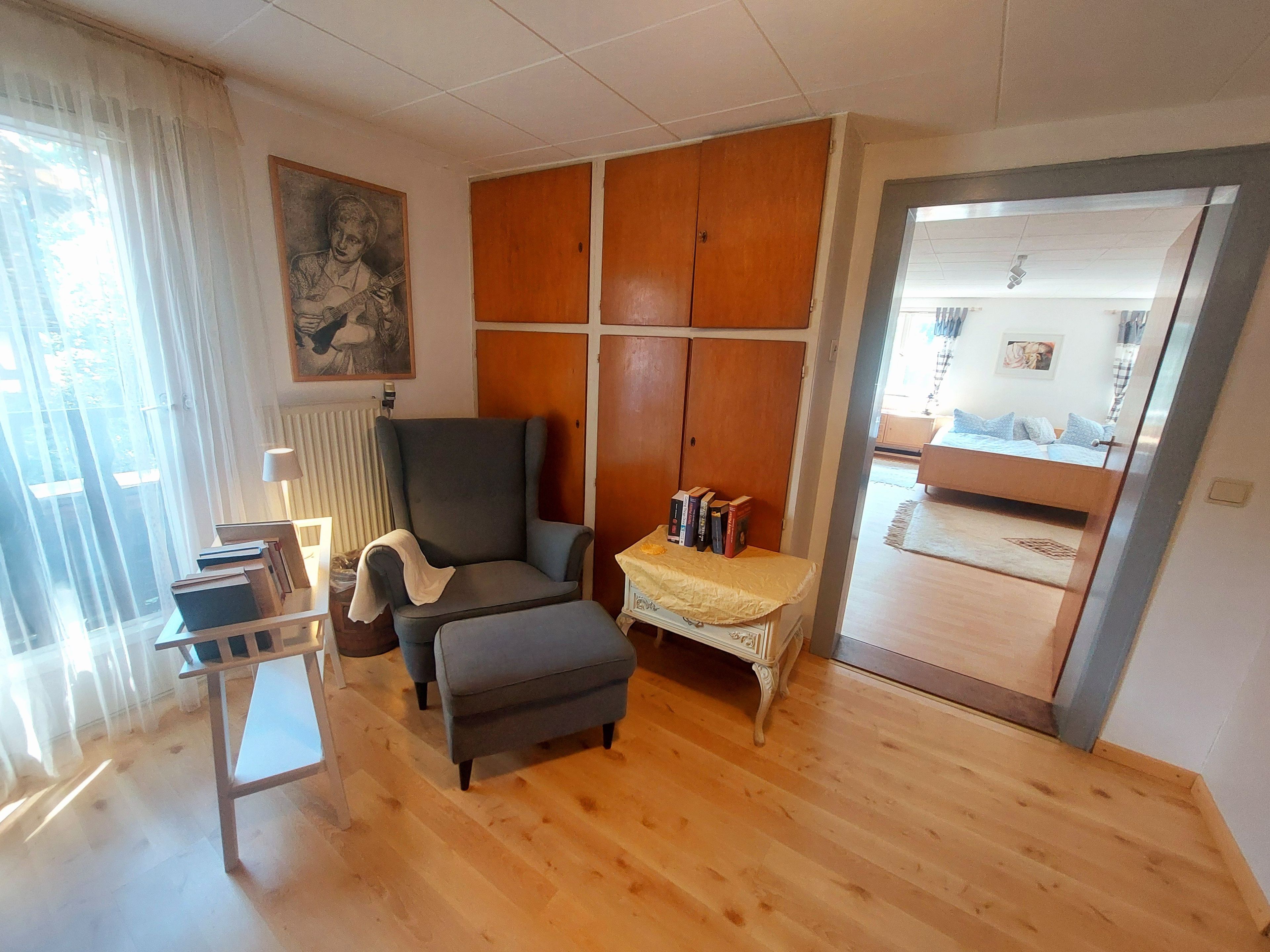 A room with a wooden floor, a grey armchair, a small table with books, a radiator, and a door leading to another room.