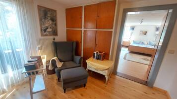 A room with a wooden floor, a grey armchair, a small table with books, a radiator, and a door leading to another room.