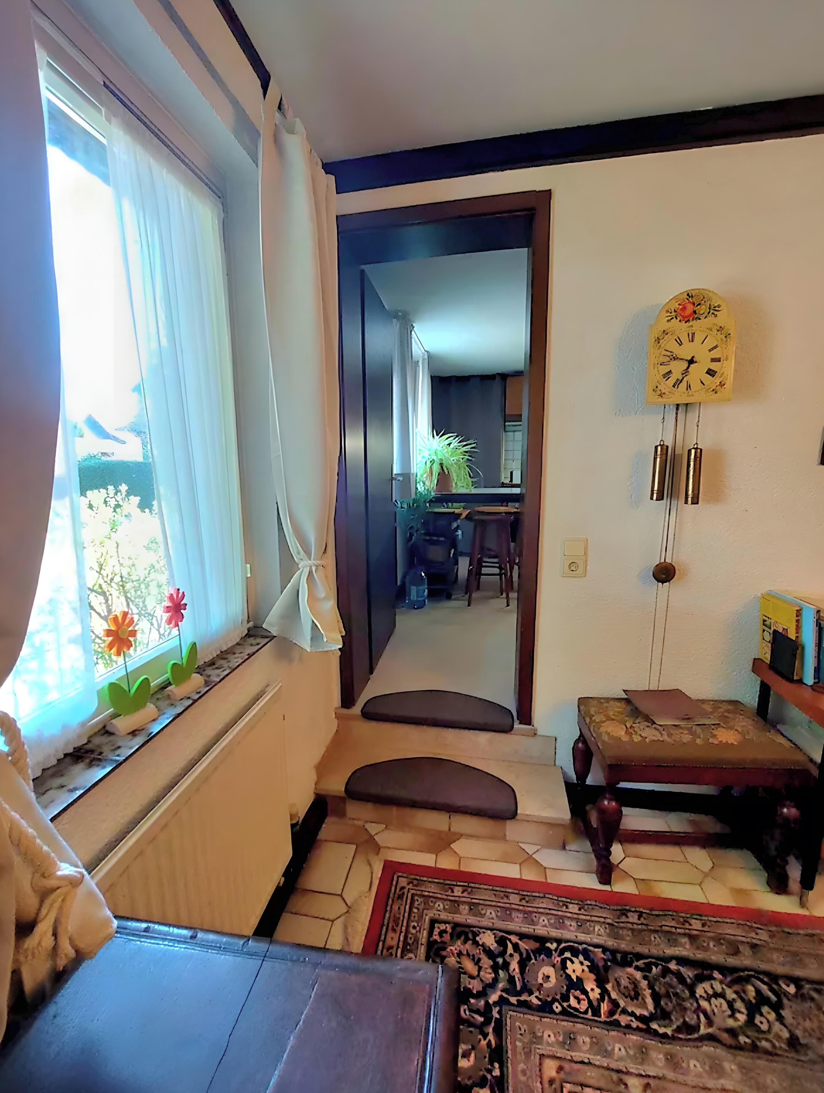 Een kamer met een raam, een klok, een plant, een bankje en een vloerkleed.
