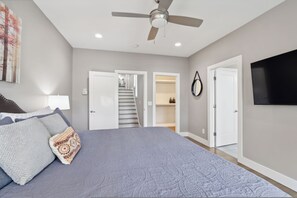 Une chambre à coucher comprenant un lit, un ventilateur de plafond, un téléviseur fixé au mur et un escalier menant à un niveau supérieur.