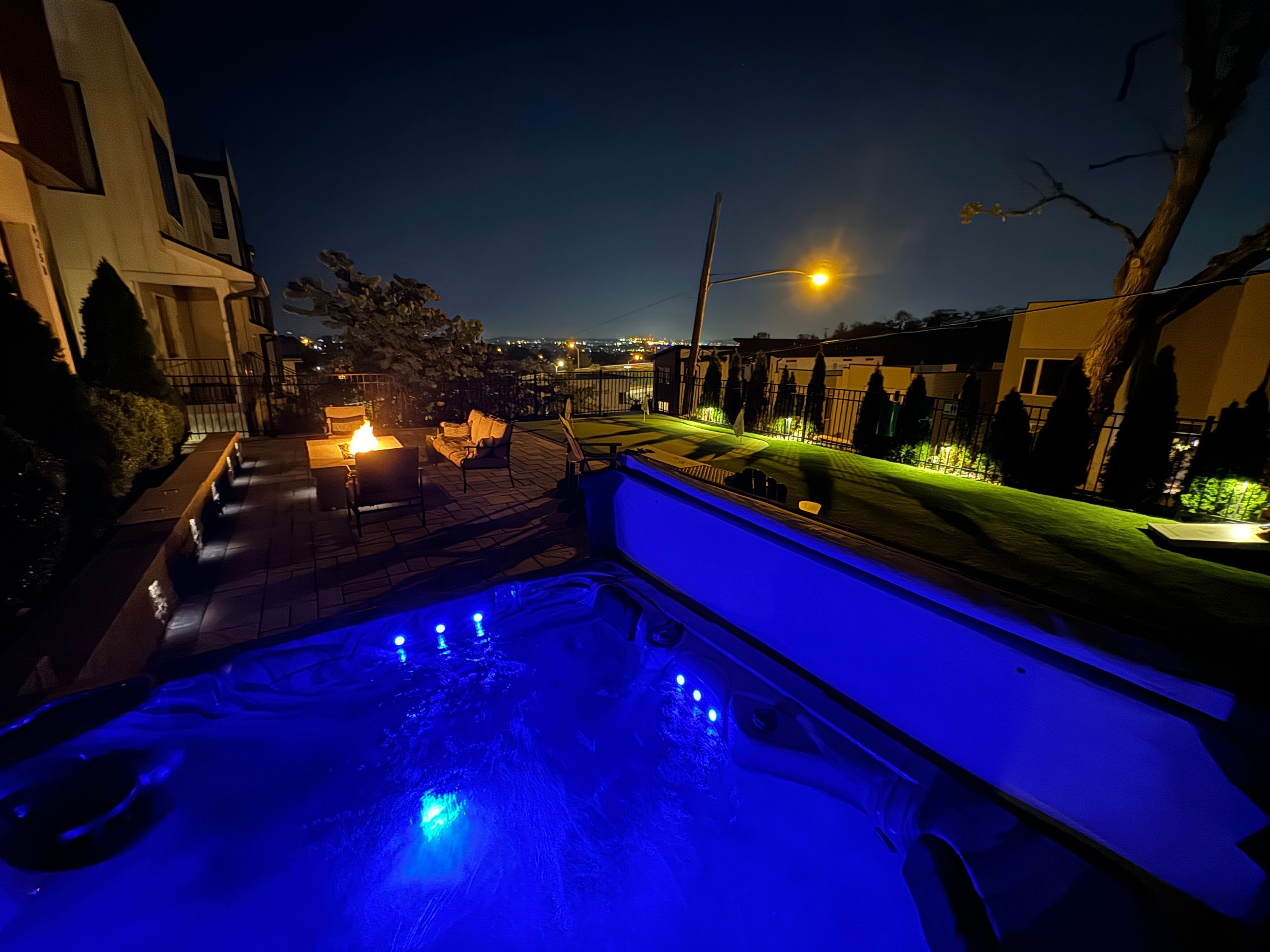 A nighttime view of a pool area with a hot tub, outdoor seating, and a well-lit pathway.