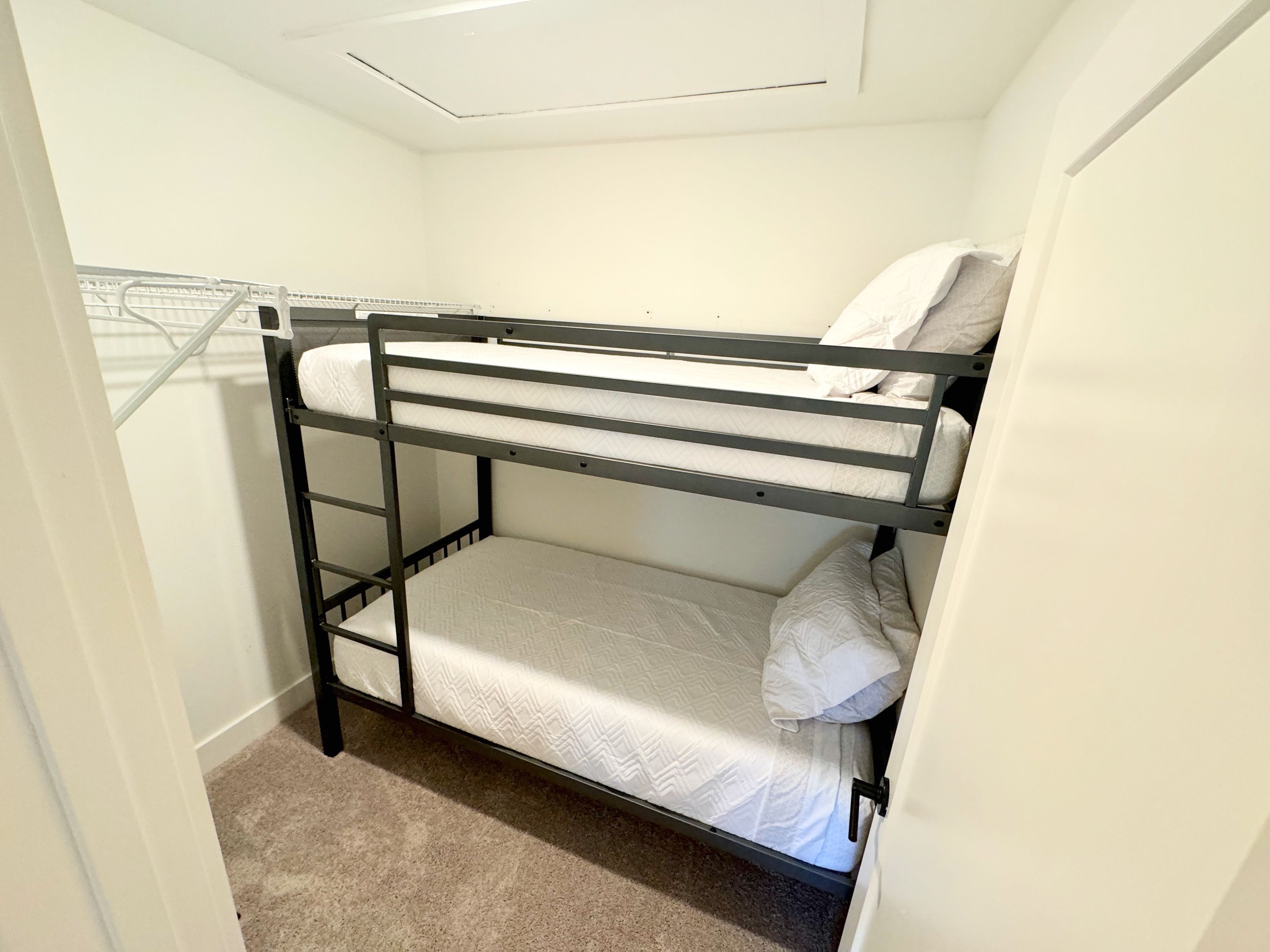 A bunk bed with a metal frame and white bedding in a small room with a carpeted floor and a white wall.