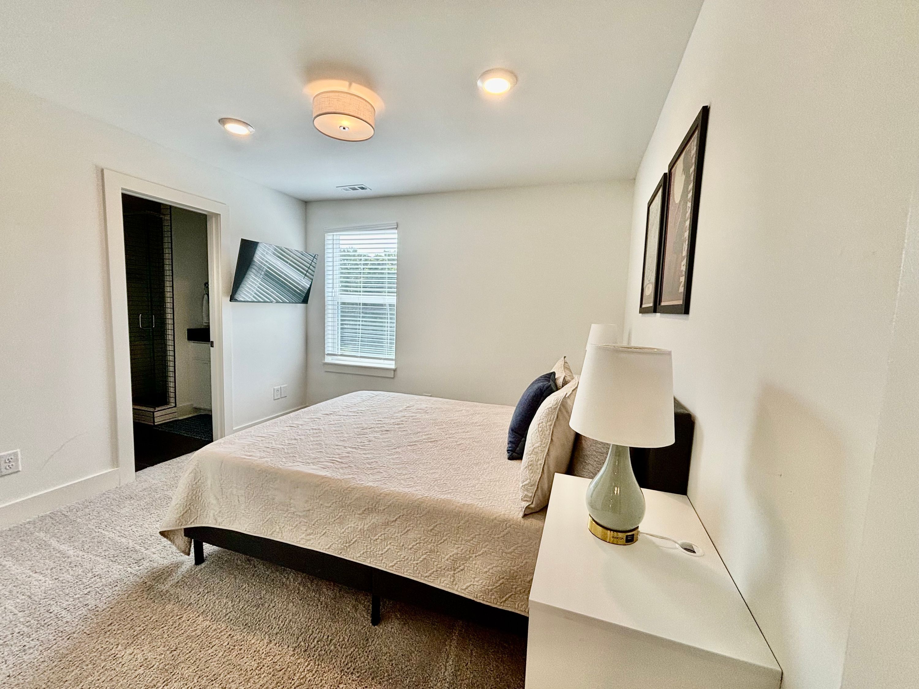 A bedroom with a bed, a bedside lamp, a window with blinds, and a door leading to a bathroom.