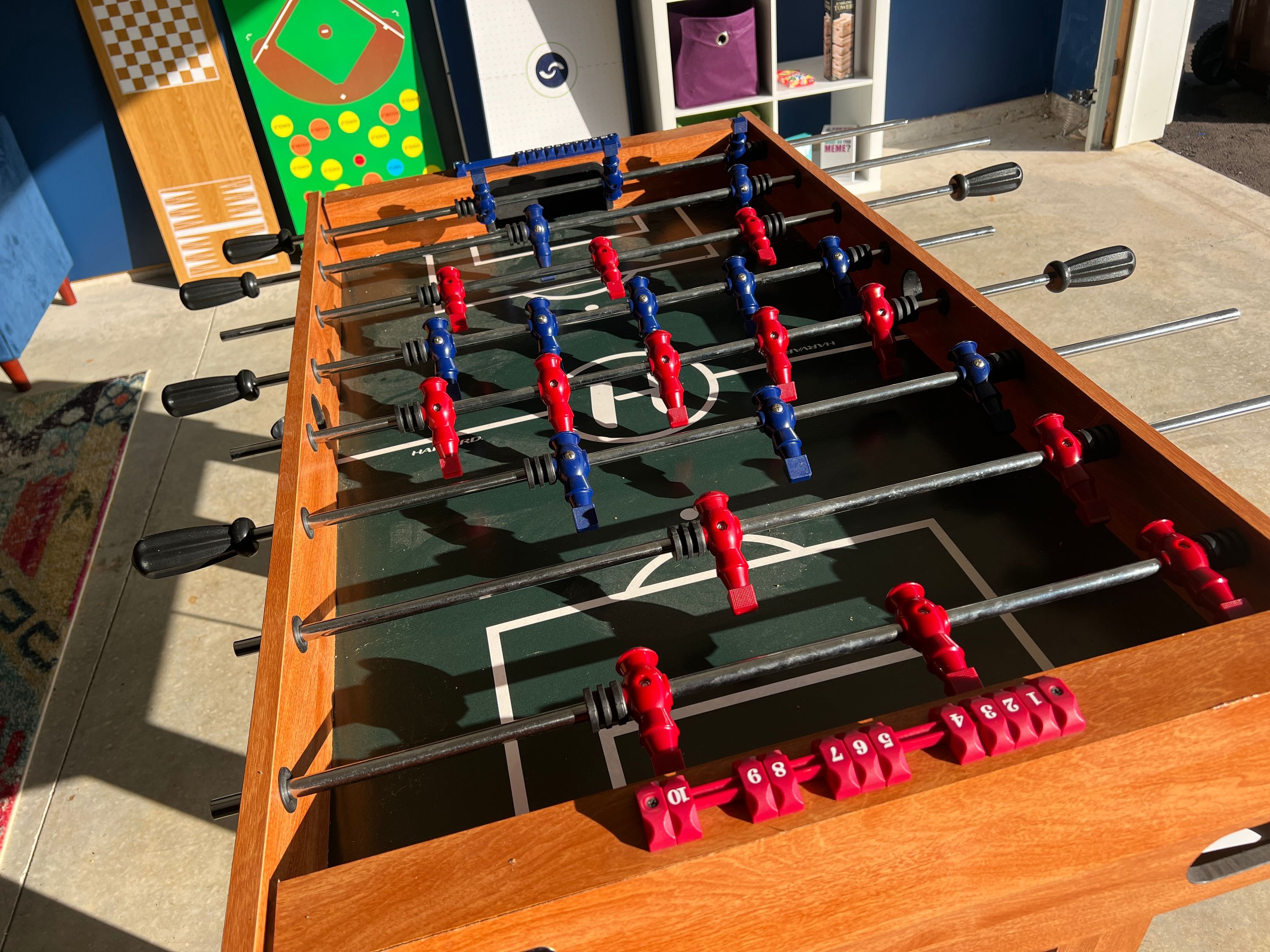 Ein Tischkicker mit roten und blauen Spielfiguren, einem Schachbrettmuster auf der Spielfläche und einem Regal mit Gegenständen im Hintergrund.