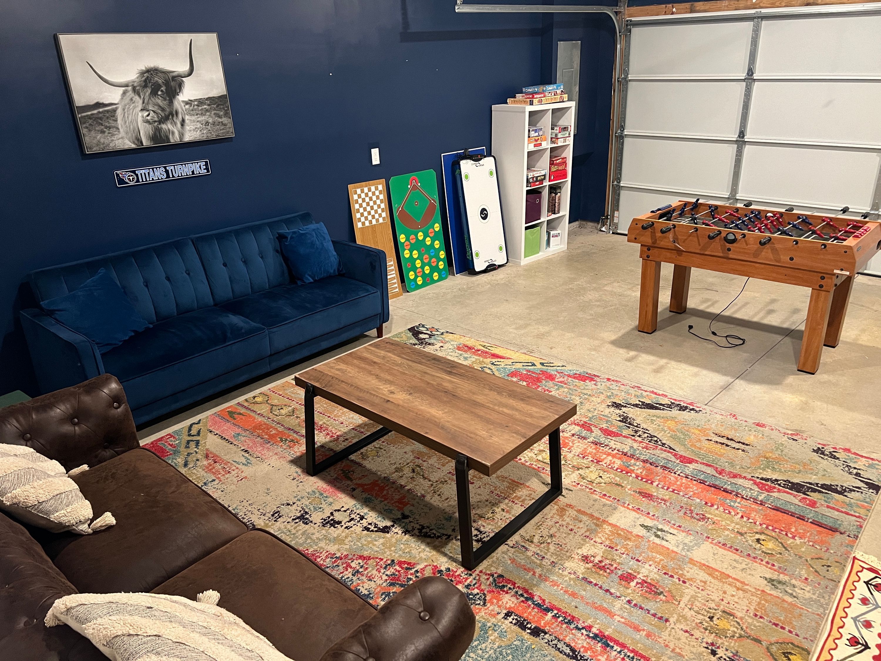 Ein Wohnzimmer mit blauem Sofa, braunem Ledersitzhocker, einem Holztisch, einem farbenfrohen Teppich, einem Kicker und einem an der Wand montierten Fernseher.