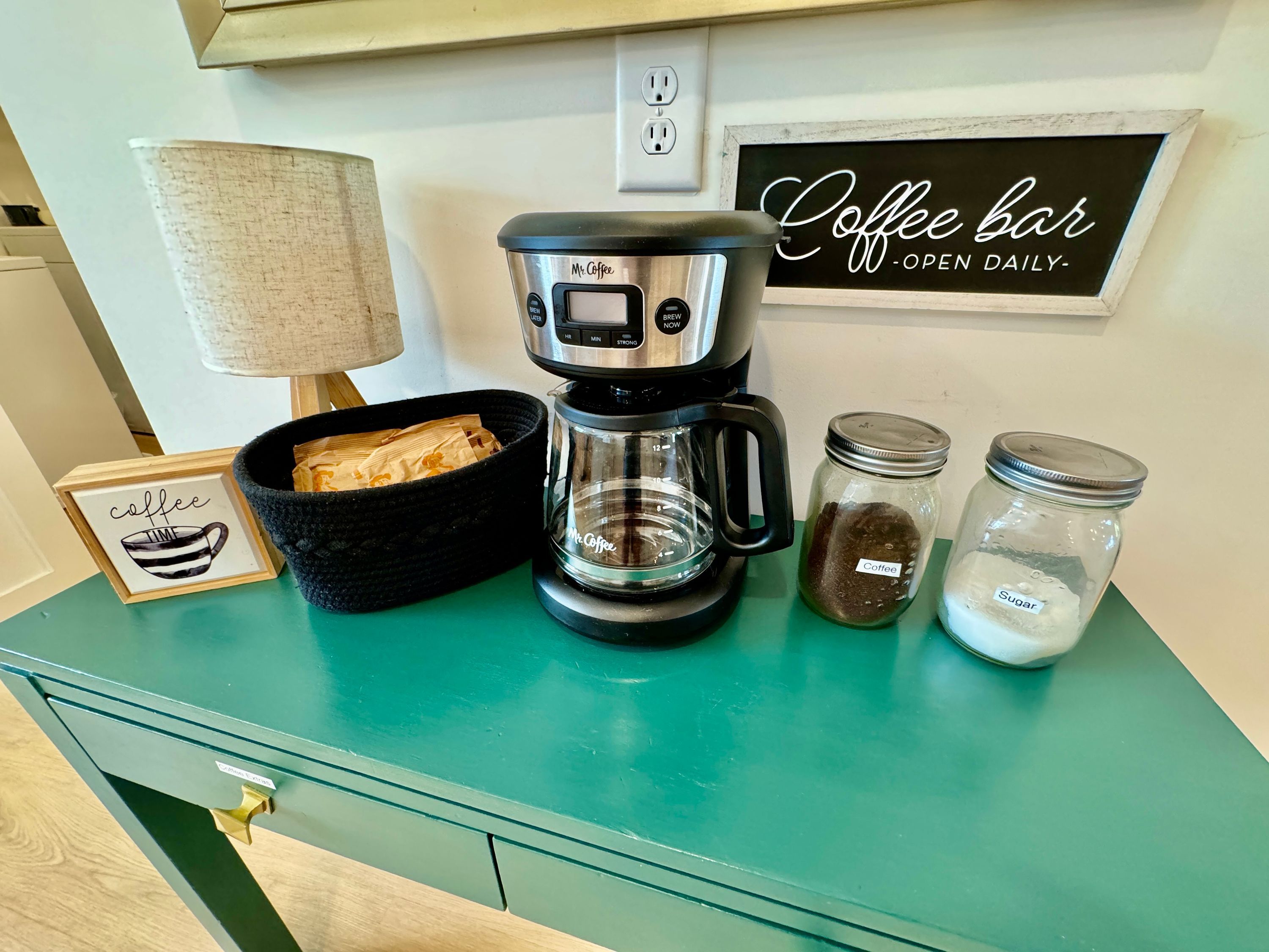 Une machine à café, un panier de dosettes de café, deux bocaux étiquetés « Café » et « Sucre », et une pancarte indiquant « Bar à café – ouvert tous les jours ».