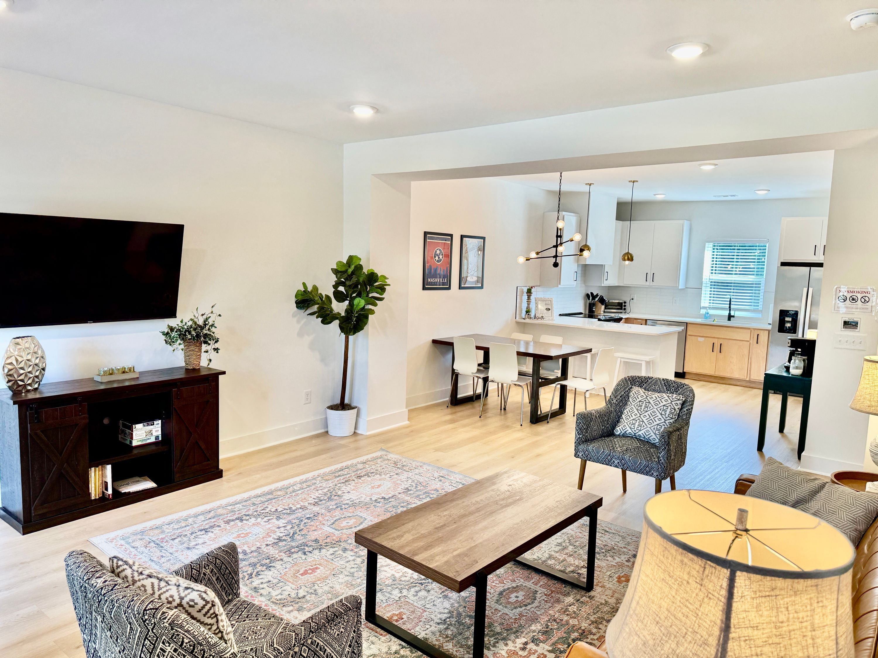 A modern living room with a flat-screen TV, a dark wooden entertainment unit, patterned sofas, a coffee table, and a dining area with a kitchen in the background.