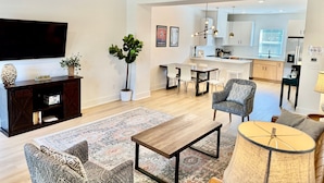 Ein modernes Wohnzimmer mit einem Flachbildfernseher, einer niedrigen Unterhaltungsecke aus dunklem Holz, gemusterten Sofas, einem Couchtisch und einem Essbereich mit einer Küche im Hintergrund.