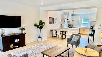 A modern living room with a flat-screen TV, a dark wooden entertainment unit, patterned sofas, a coffee table, and a dining area with a kitchen in the background.