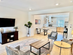 A modern living room with a flat-screen TV, a dark wooden entertainment unit, patterned sofas, a coffee table, and a dining area with a kitchen in the background. - Closetobroadwayairportoprymillsgameroom (Nashville)