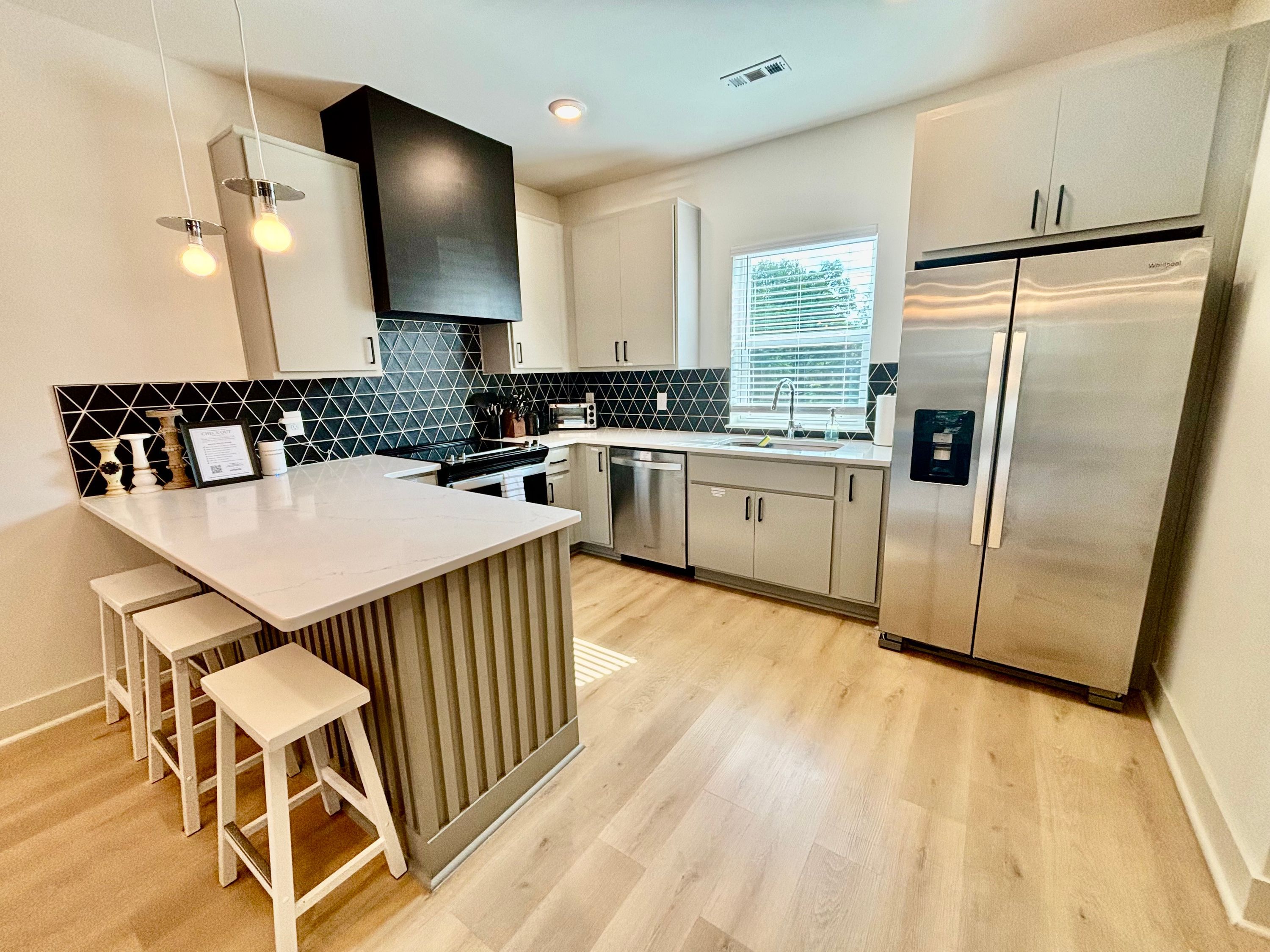 Et moderne køkken med en central ø, barstole, et rustfrit stålkøleskab og en sort-hvid backsplash.