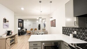 A modern kitchen with a central island, built-in oven, and microwave. There is a dining area with a table and chairs, and a coffee bar with a sign. - Closetobroadwayairportoprymillsgameroom12 (Nashville)