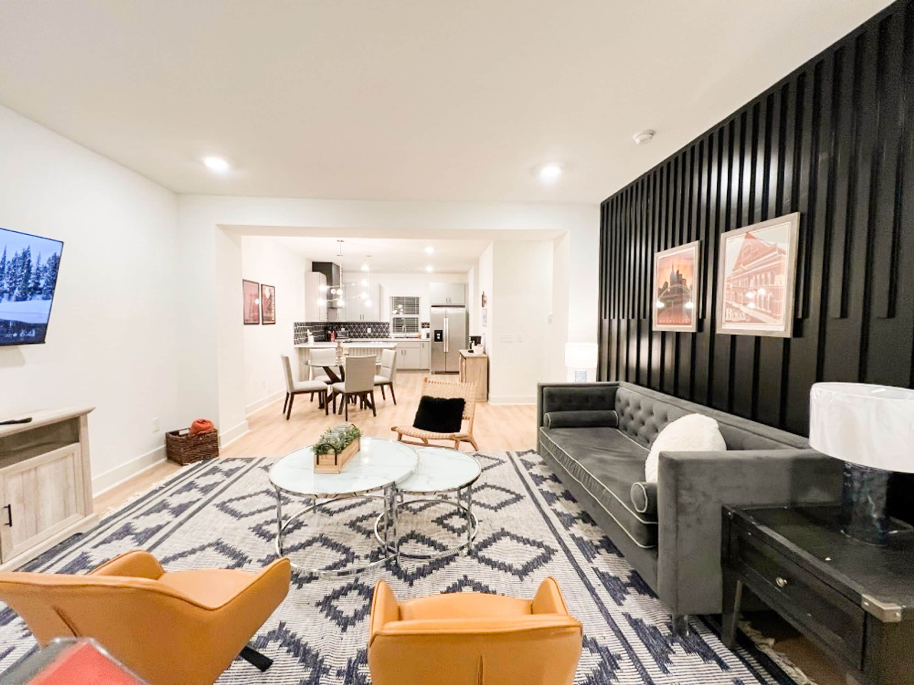 Una sala de estar moderna con un sofá, dos sillones, una mesa de centro y un televisor. Se observa una cocina al fondo.