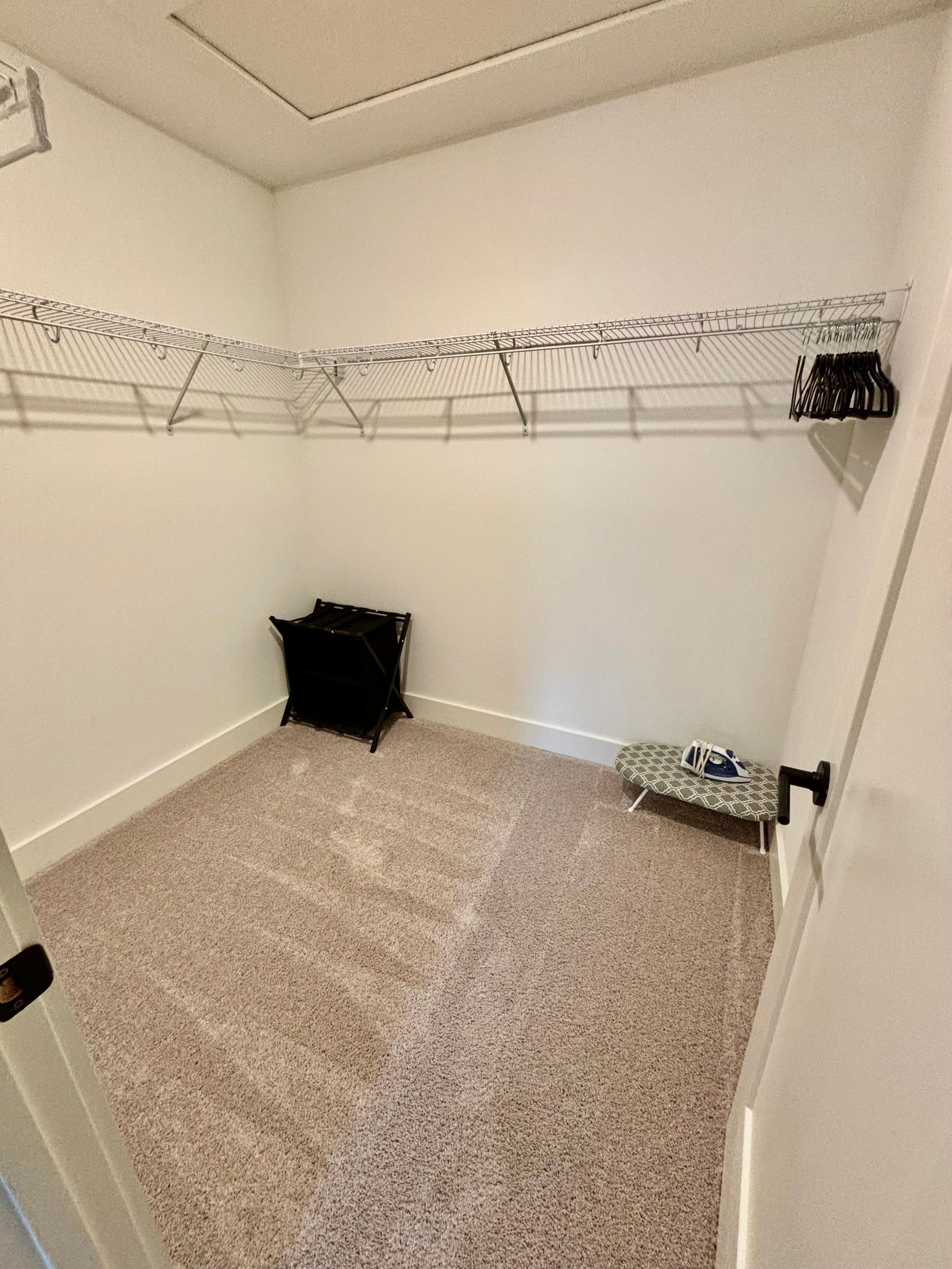 Quarto pequeno com carpete, um varal de metal, uma unidade de armazenamento preta e um espaço para passar roupa.
