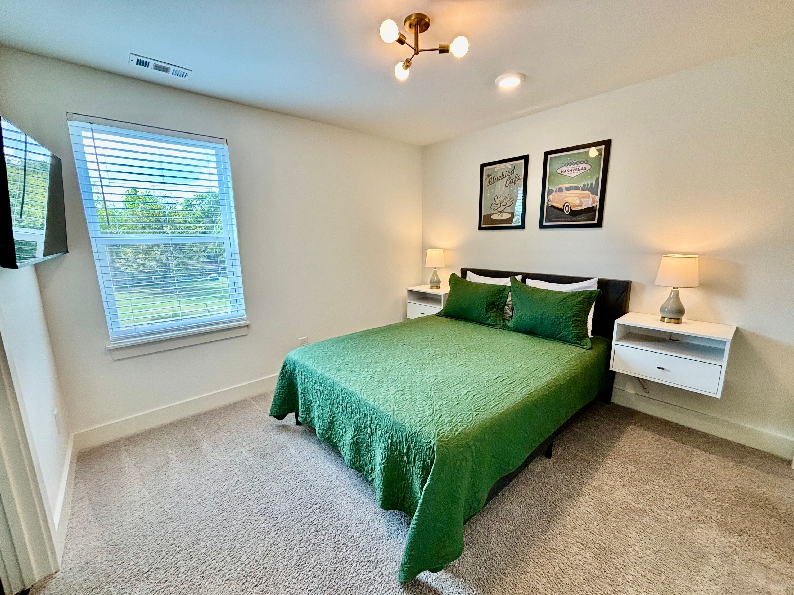 A bedroom with a green bedspread, two bedside tables with lamps, a window with blinds, and two framed pictures on the wall.
