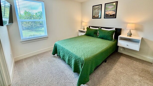 A bedroom with a green bedspread, two bedside tables with lamps, a window with blinds, and two framed pictures on the wall. - Closetobroadwayairportoprymillsgameroom12 (Nashville)