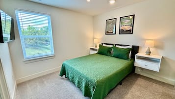 A bedroom with a green bedspread, two bedside tables with lamps, a window with blinds, and two framed pictures on the wall.