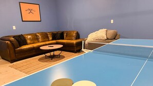 A room with a blue ping pong table, two ping pong paddles, a brown leather sofa, a small round table, and a framed picture on the wall. - Closetobroadwayairportoprymillsgameroom12 (Nashville)