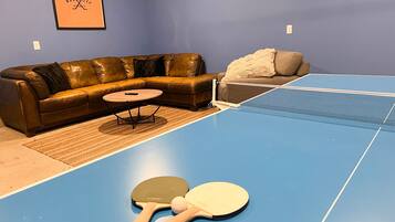 A room with a blue ping pong table, two ping pong paddles, a brown leather sofa, a small round table, and a framed picture on the wall.