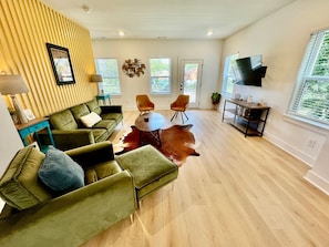 A spacious living room with a green sofa set, two orange chairs, a round coffee table, and a large leather rug. There is a flat-screen TV mounted on the wall and a wooden cabinet with decorative items. - Closetobroadwayairportoprymillsgameroom (Nashville)