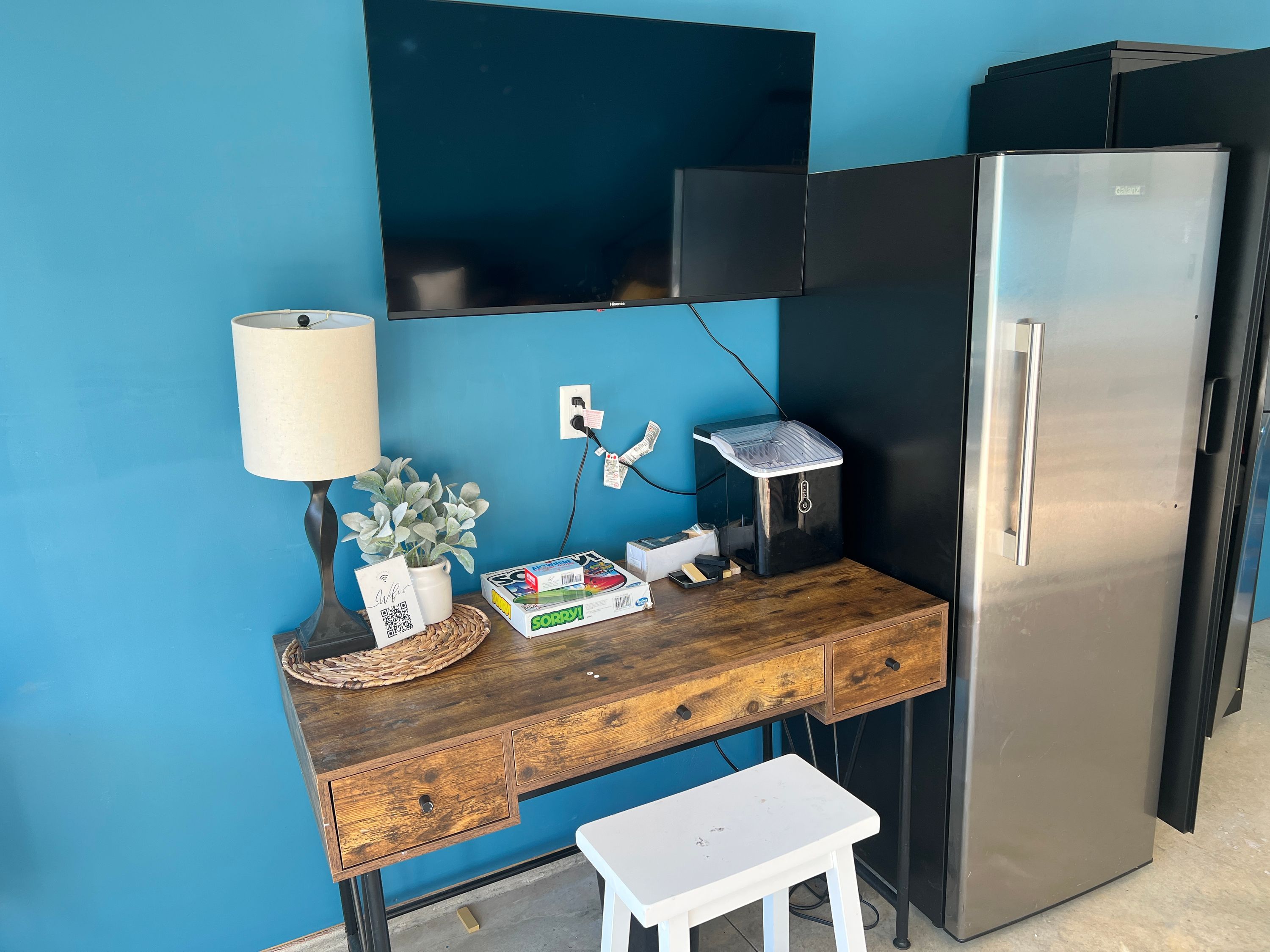 Een kitchenette met een roestvrijstalen koelkast, een houten bureau met een lamp, en een aan de muur bevestigde televisie.