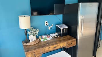 Een kitchenette met een roestvrijstalen koelkast, een houten bureau met een lamp, en een aan de muur bevestigde televisie.