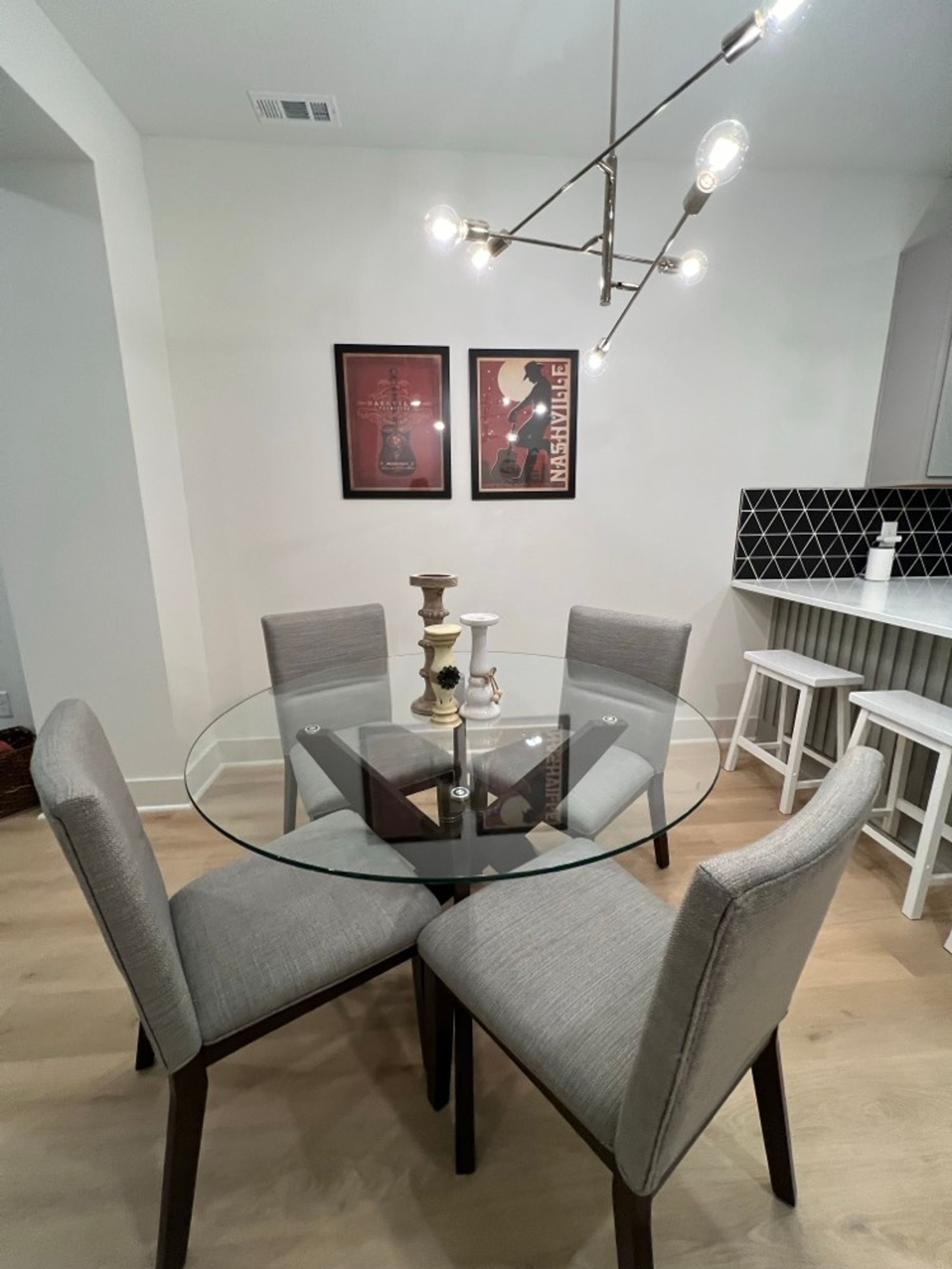 Un comedor moderno con una mesa de vidrio rodeada por cuatro sillas tapizadas en gris. Hay dos carteles enmarcados en la pared, una lámpara colgante sobre la mesa y una encimera blanca con un salpicadero de rejilla negra al fondo.