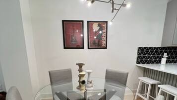 Un comedor moderno con una mesa de vidrio rodeada por cuatro sillas tapizadas en gris. Hay dos carteles enmarcados en la pared, una lámpara colgante sobre la mesa y una encimera blanca con un salpicadero de rejilla negra al fondo.