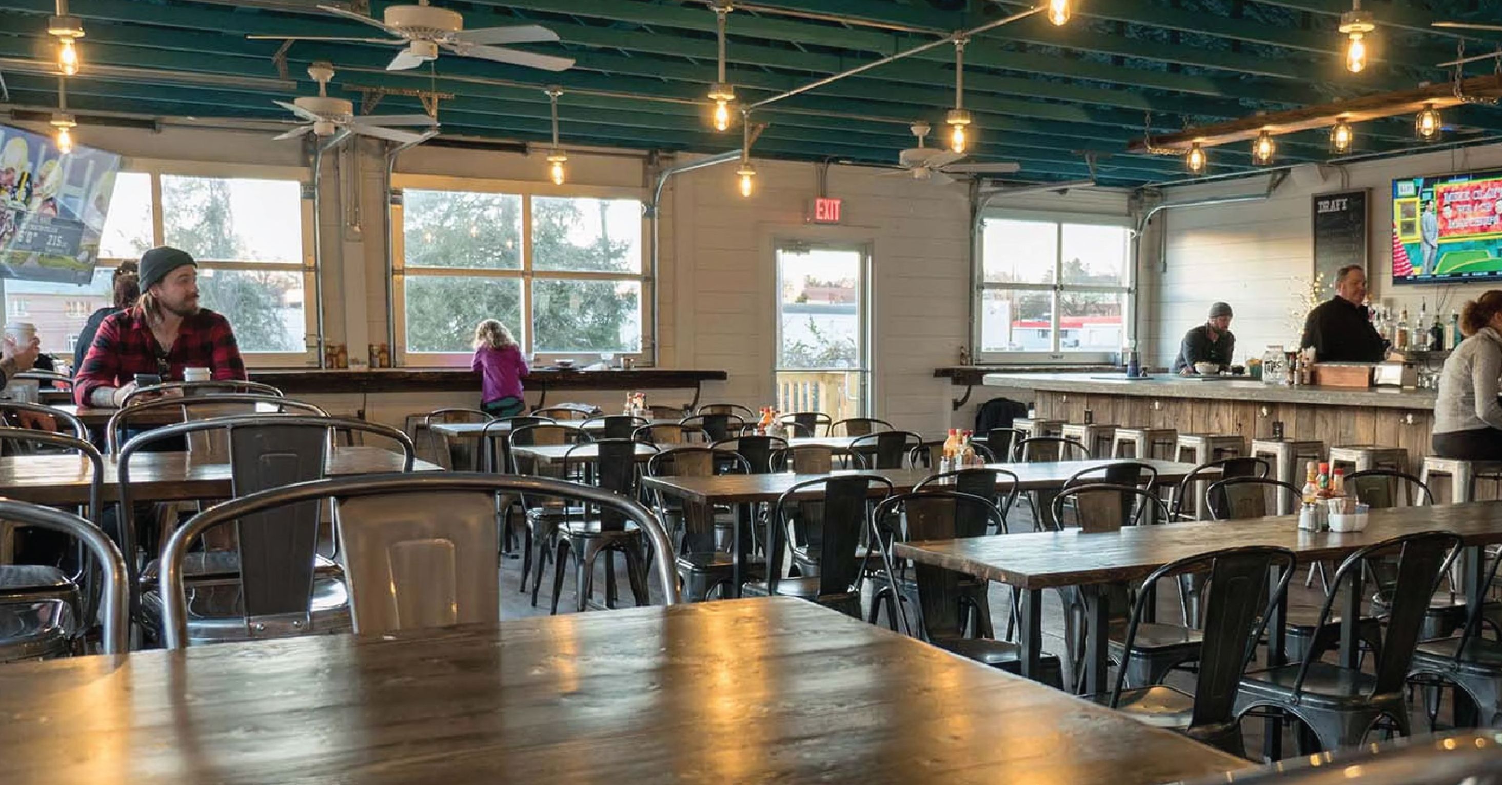 A restaurant interior with multiple tables and chairs, ceiling fans, and large windows.