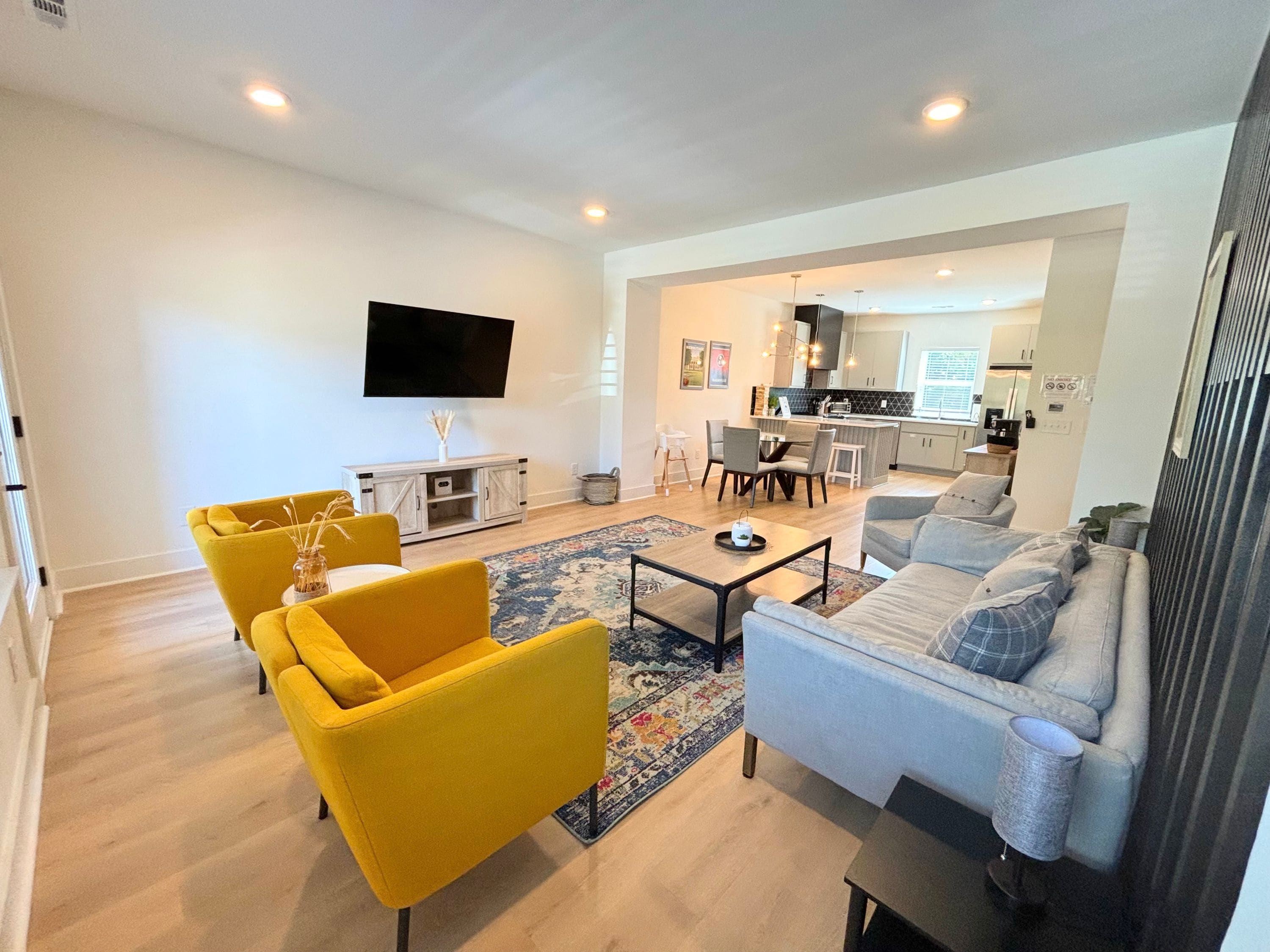 A modern living room with a sofa, armchairs, and a coffee table. There is a flat-screen TV mounted on the wall and a kitchen area visible in the background.