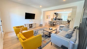 A modern living room with a sofa, armchairs, and a coffee table. There is a flat-screen TV mounted on the wall and a kitchen area visible in the background.