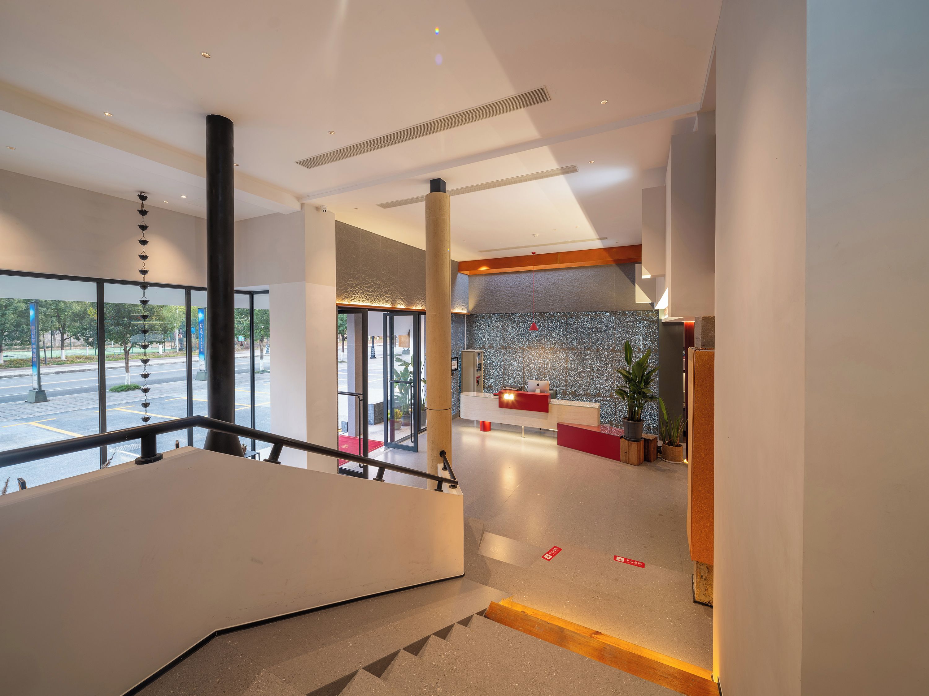 A modern hotel lobby with a reception desk, a staircase, and a view of the street outside.