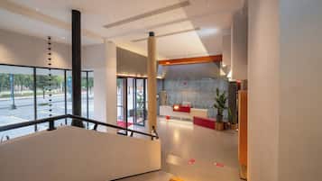 A modern hotel lobby with a reception desk, a staircase, and a view of the street outside.