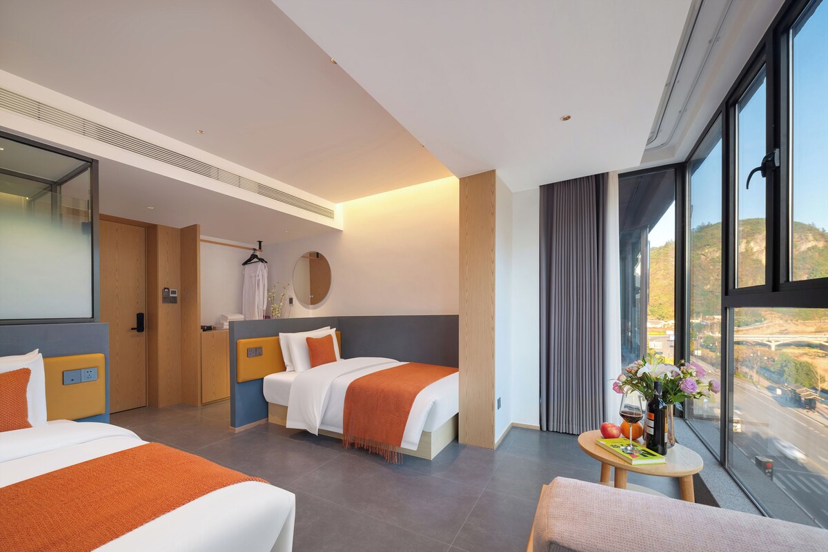 Habitación de hotel con dos camas, una zona de estar, una mesa redonda con un jarrón de flores y un ventanal con vistas.