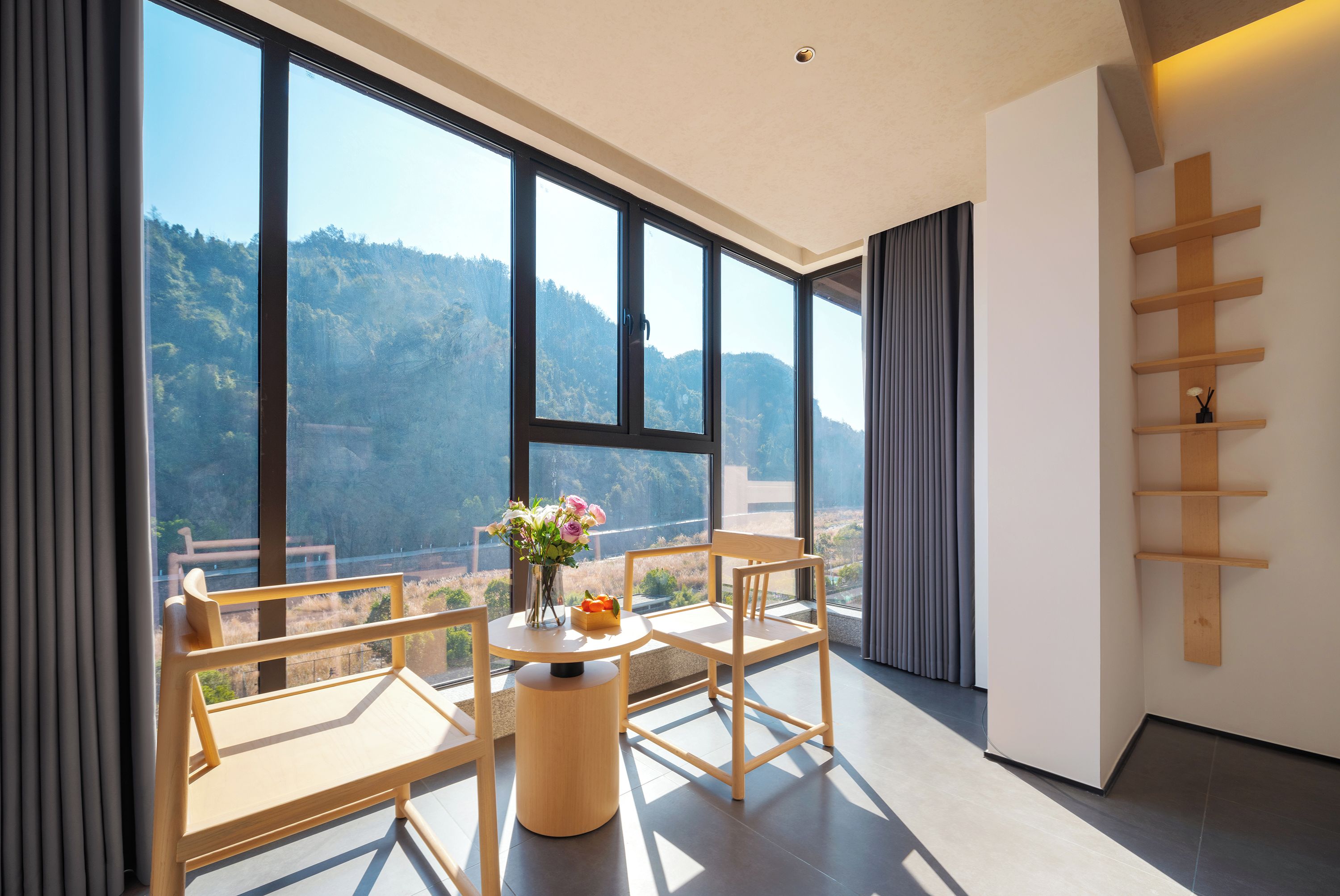 Une pièce moderne avec de grandes fenêtres, une table ronde, deux chaises, un vase avec des fleurs et une étagère en bois.