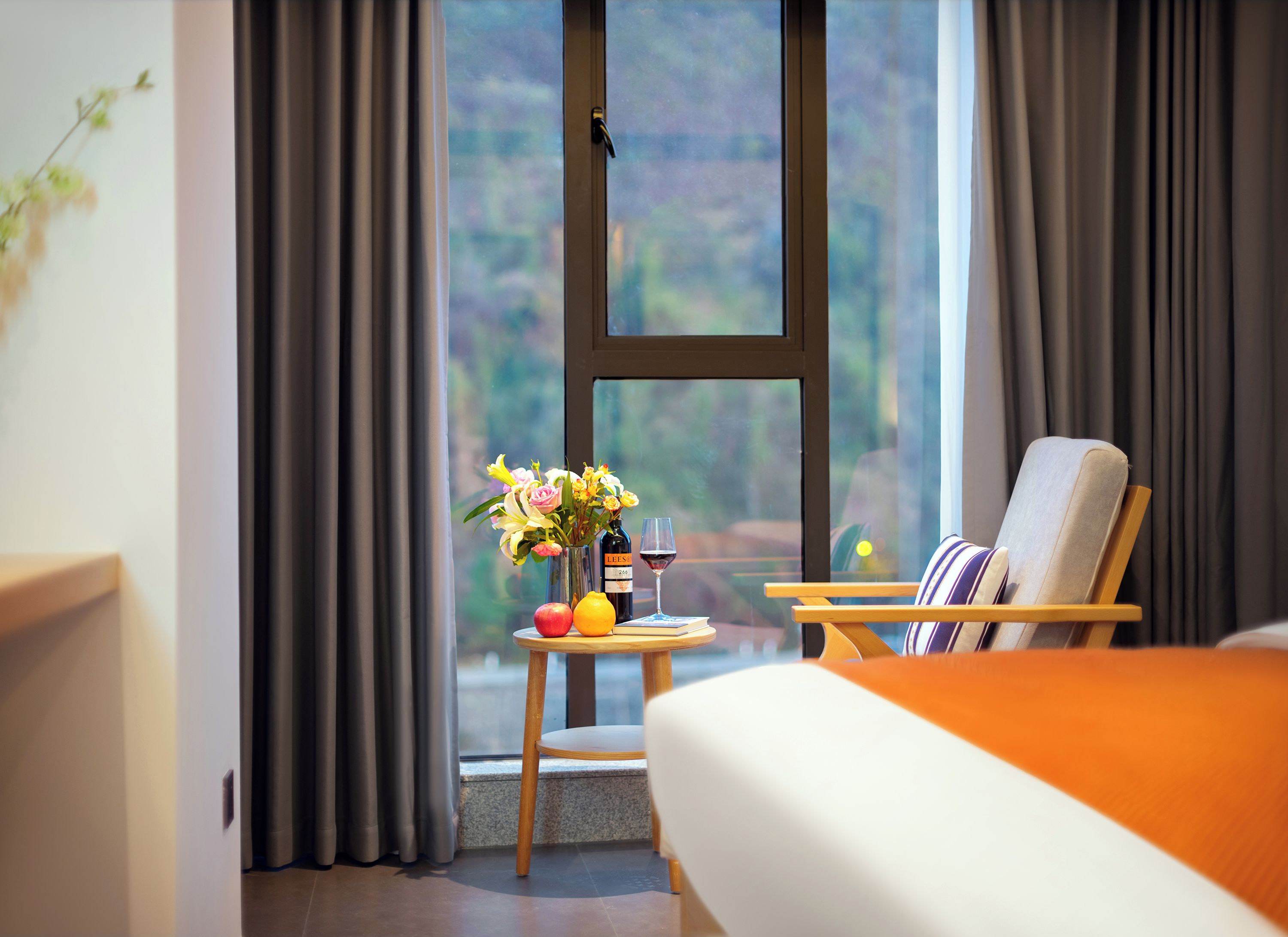 A hotel room with a bed, a chair, a small table with a vase of flowers, a bottle, and a glass of red wine.