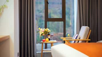 A hotel room with a bed, a chair, a small table with a vase of flowers, a bottle, and a glass of red wine.
