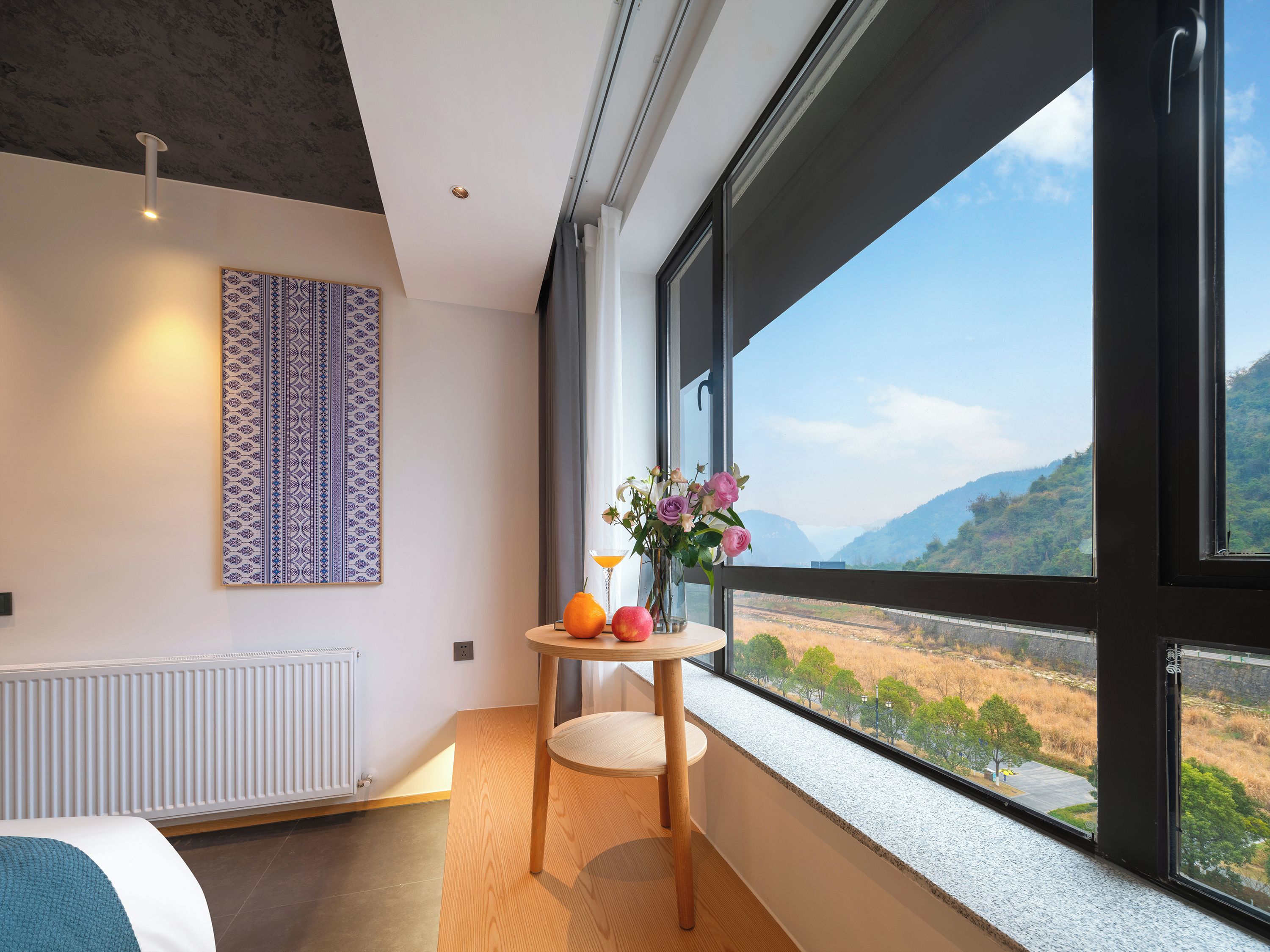 Une chambre moderne avec une grande fenêtre, une table ornée de fruits et de fleurs, un radiateur et un mur à motifs.