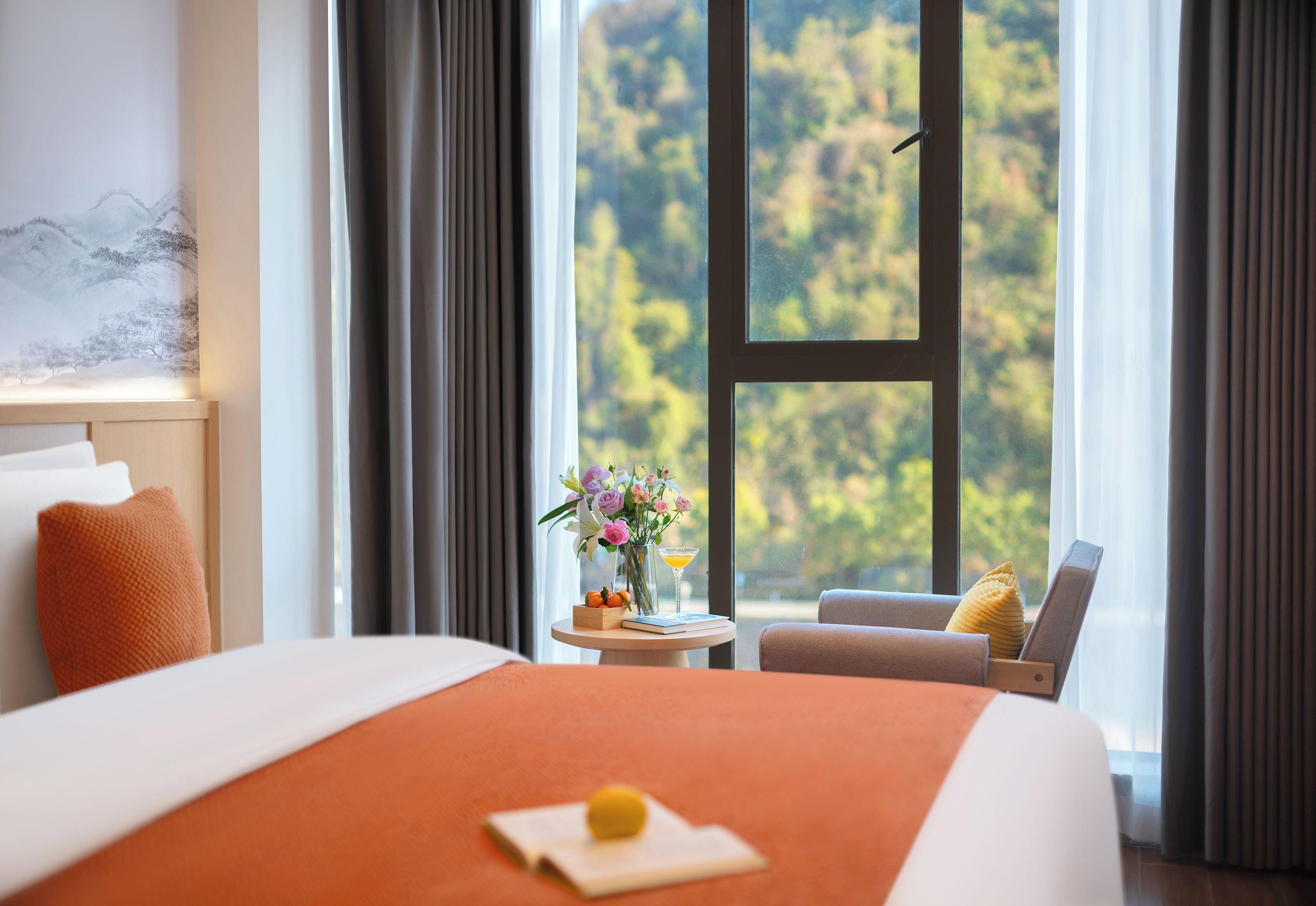 A hotel room with a large bed, a small round table with a vase of flowers, a chair, and a view of trees through a window.