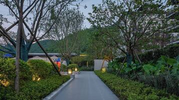 A pathway flanked by lush greenery and trees, leading to a building with a traditional roof.