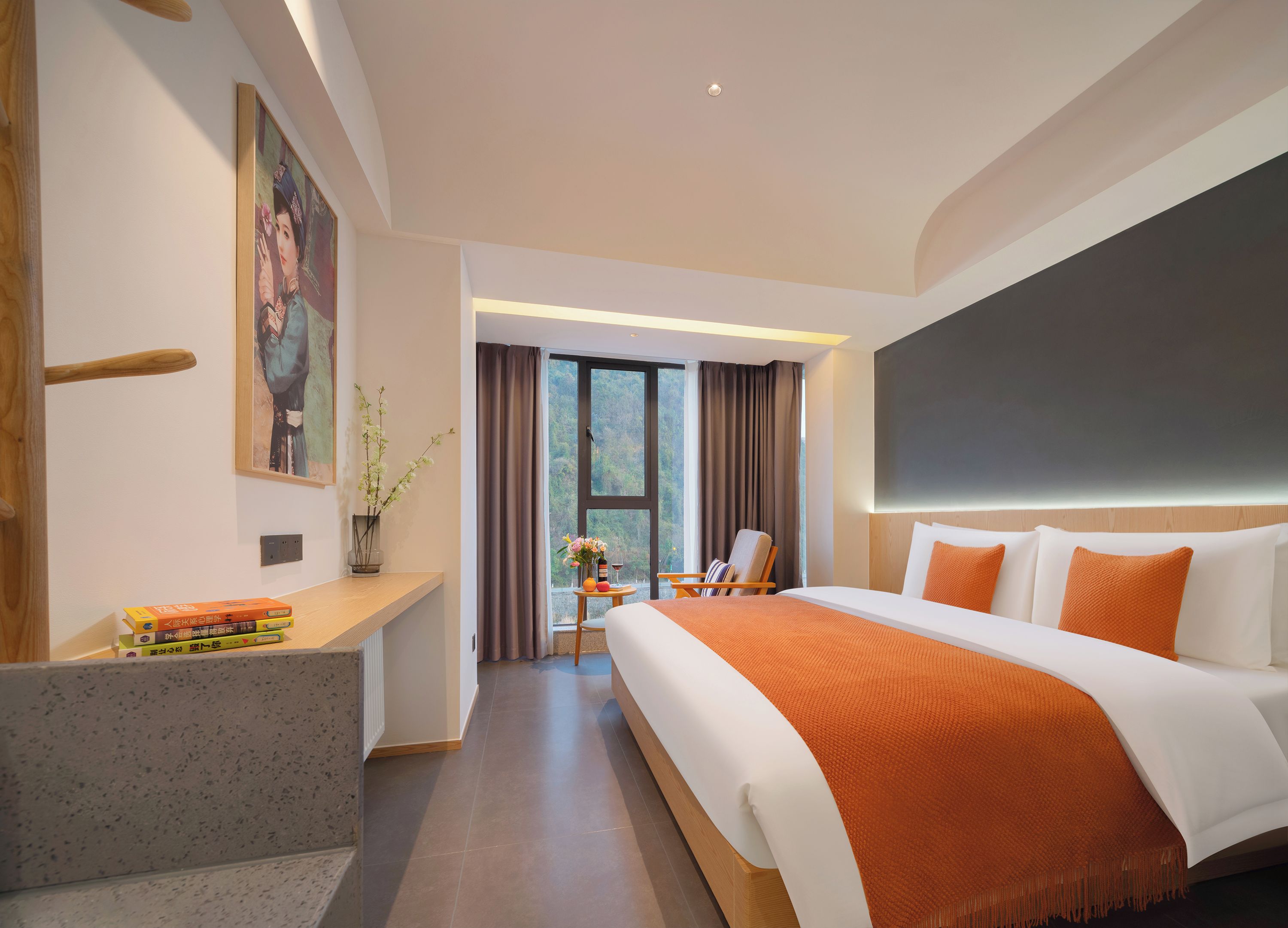 Quarto de hotel moderno com uma cama grande, uma escrivaninha com livros, uma cadeira e vista para o exterior.
