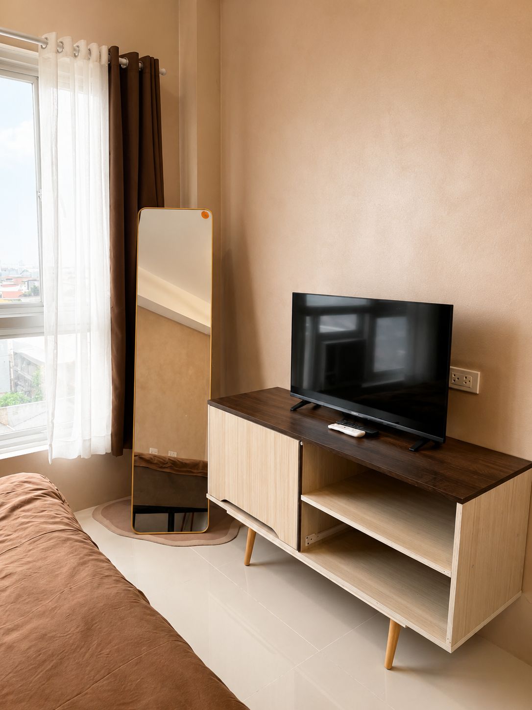 Una moderna camera d'albergo con una TV a schermo piatto su un mobile in legno, un grande specchio e una finestra con tende.